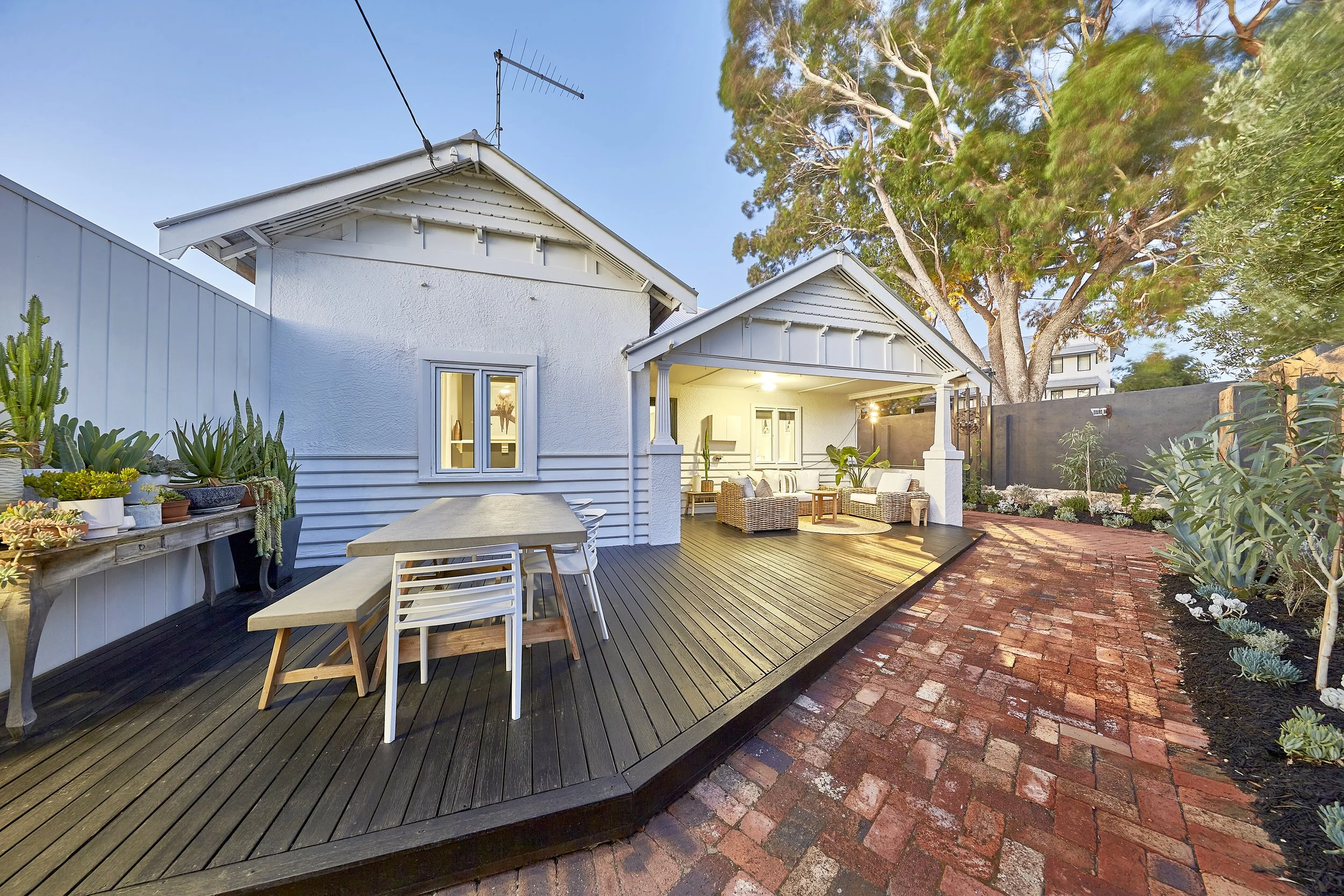 A backyard patio area with a wooden deck and brick pathway, outdoor furniture including a table and chairs, surrounded by various plants and trees, with a white house and an outdoor sofa visible in the background.