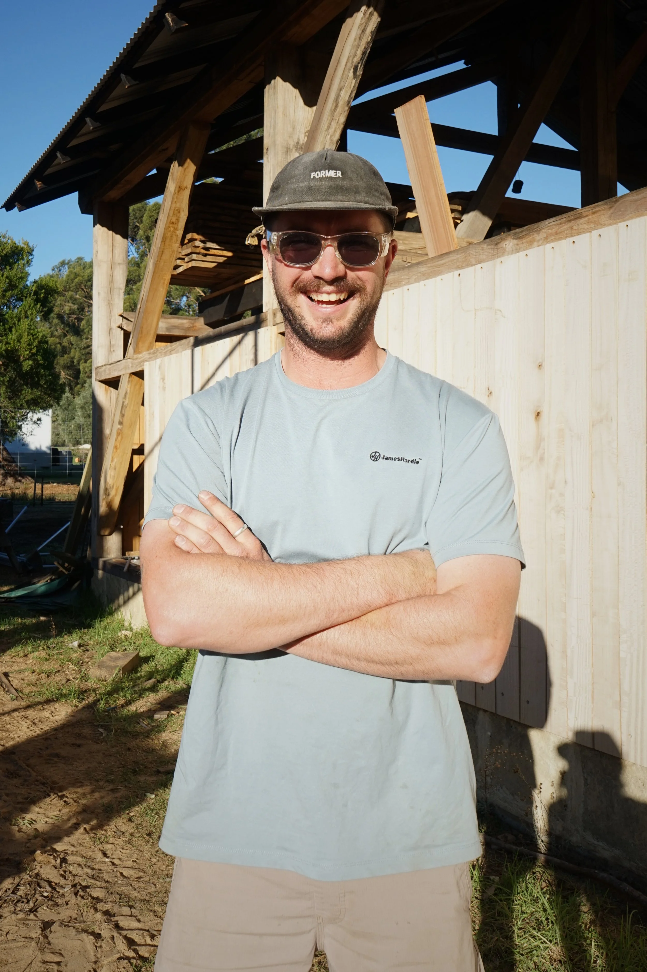 A man smiling outdoors wearing glasses, a cap with the word 'FORMER' on it, a light gray T-shirt, and beige shorts, standing in front of a wooden structure under construction.