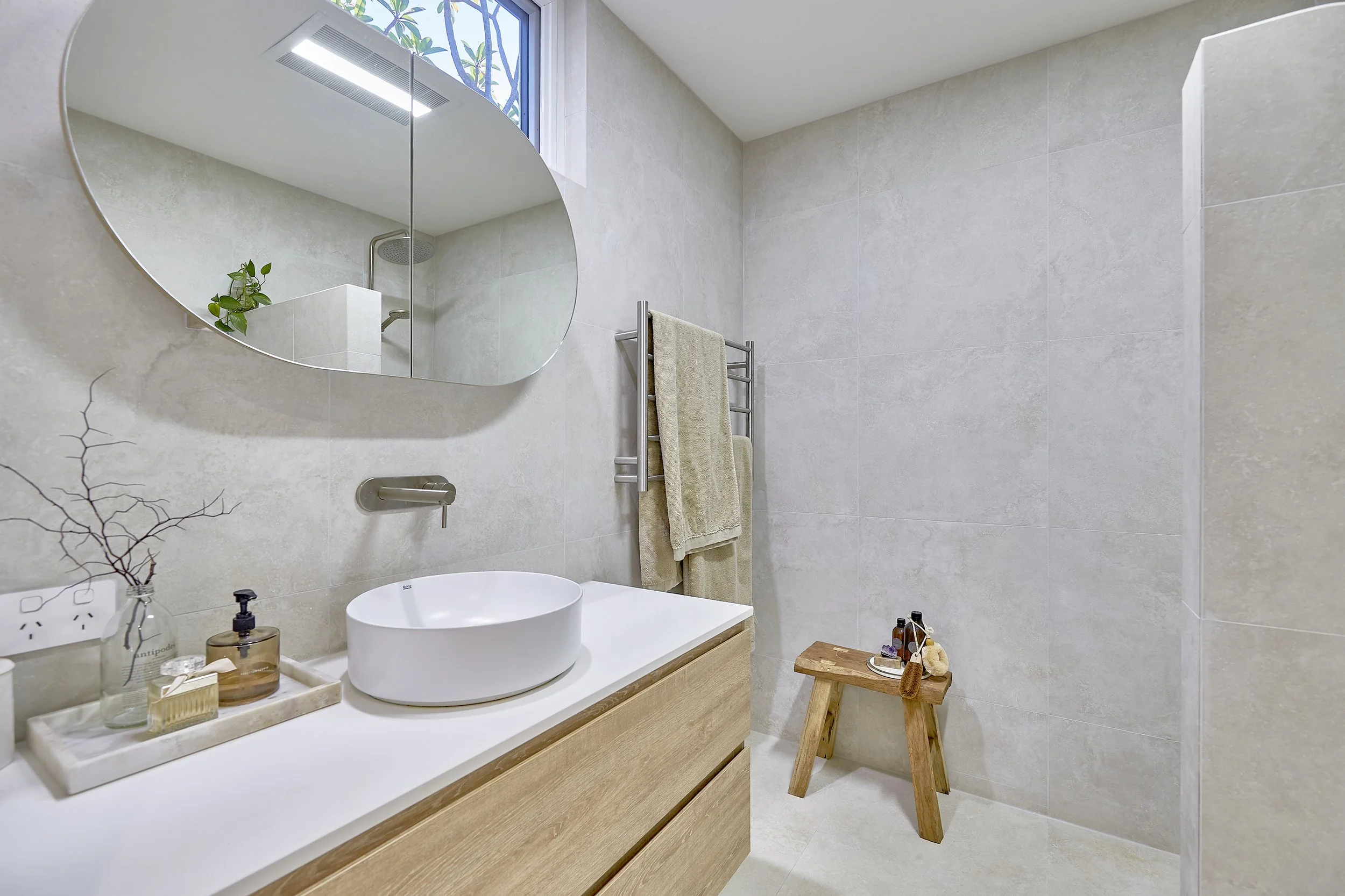 A modern, minimalist bathroom with a large oval mirror above a wooden vanity with a round vessel sink, decorated with hand soap, a glass bottle, and a tray. There is a towel on a towel rack near a window, and a small wooden stool with bath essentials
