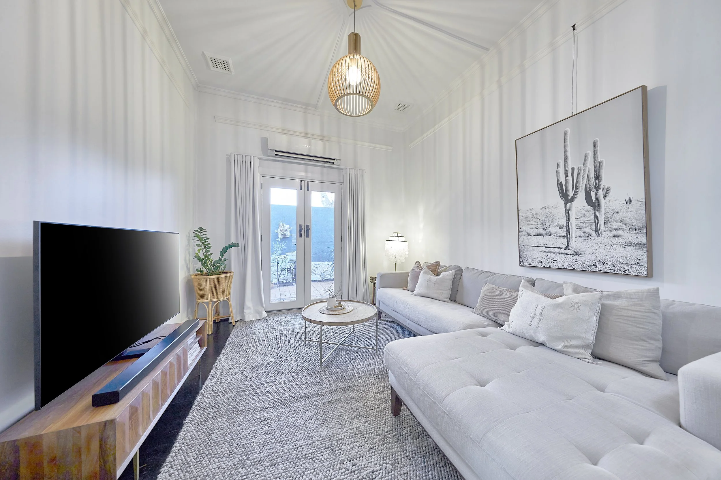 Living room with a white sofa, coffee table, large wall art of cacti, TV, potted plant, and glass door leading outside, with ceiling light fixture and curtains.