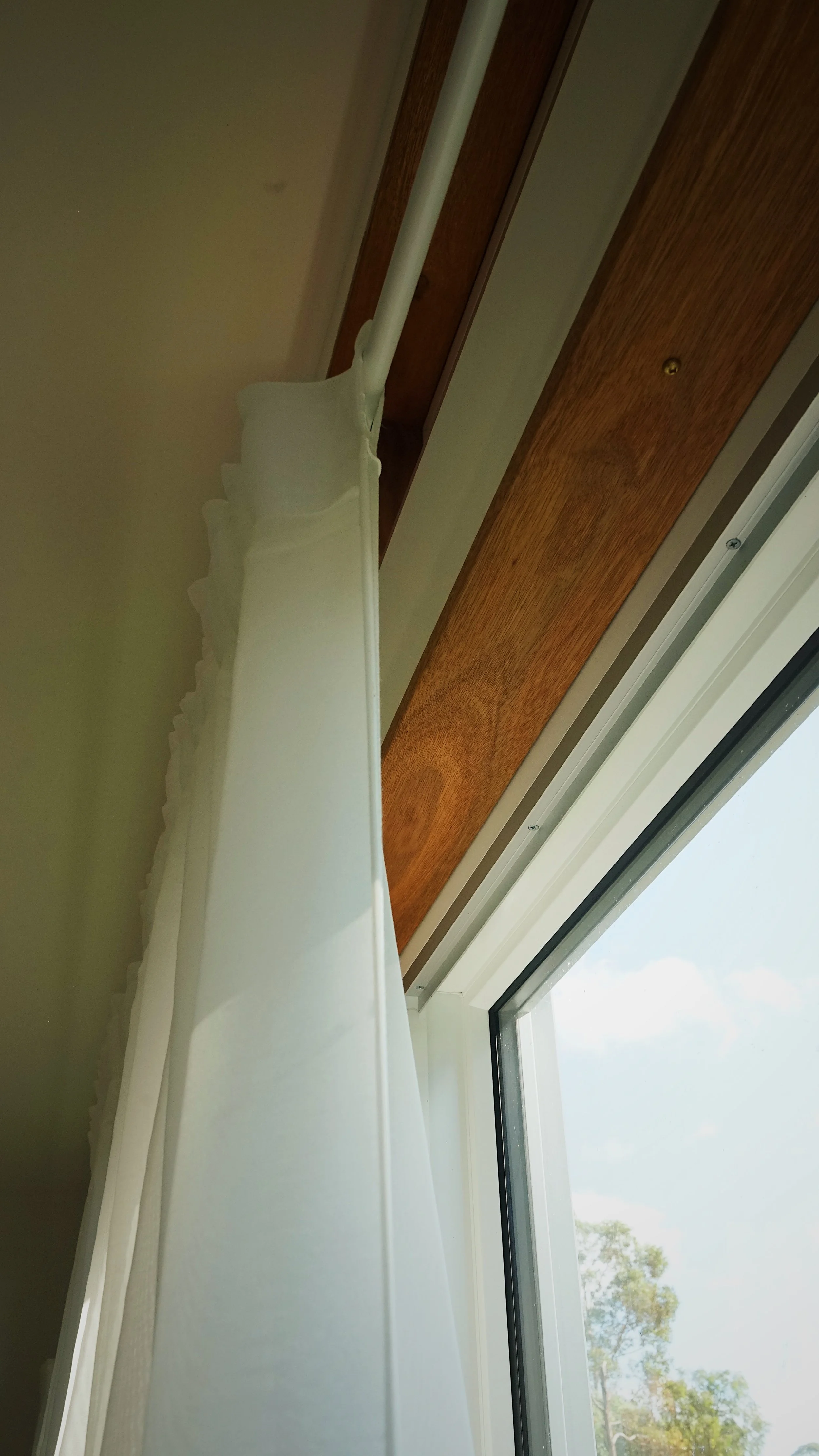Looking up at a window with a white curtain and wooden trim, outside trees are visible on a sunny day.