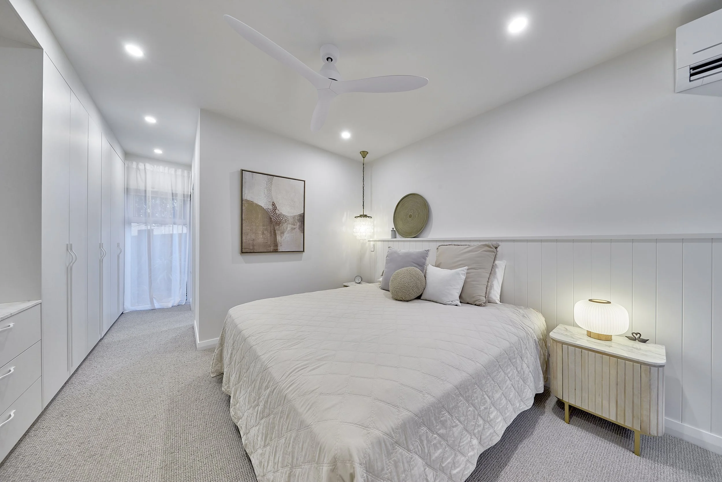 A cozy bedroom with a white bed, plush pillows, a bedside lamp, and minimalist decor. Ceiling fan and built-in wardrobe with handles are visible, along with soft natural light through a sheer curtain.