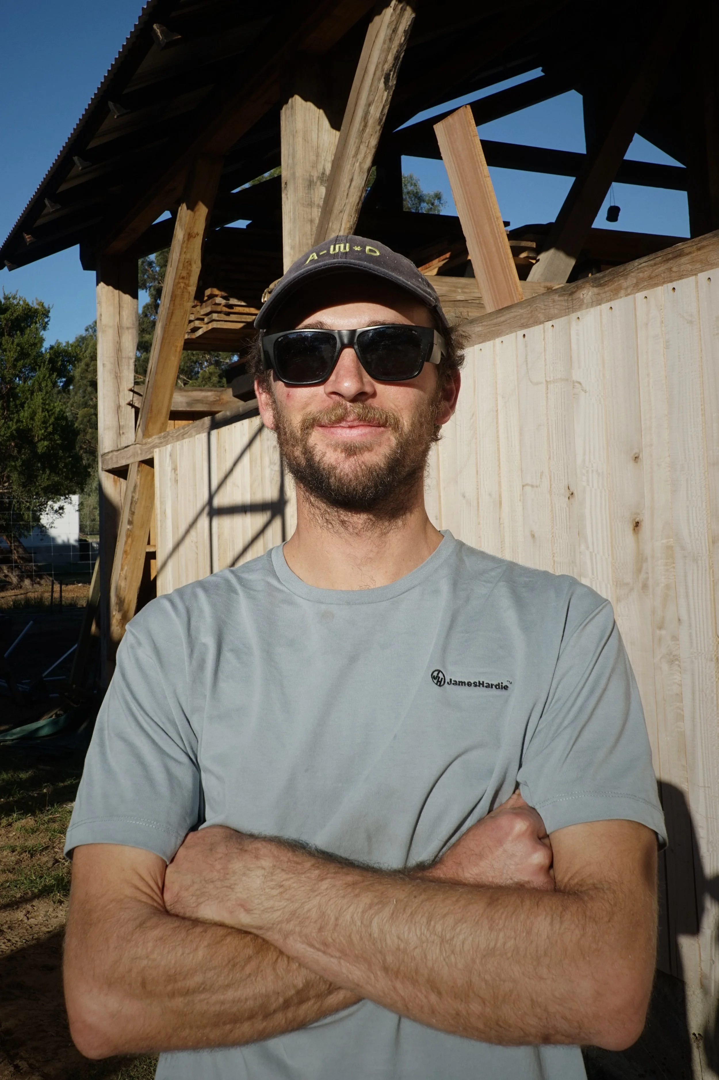 A man with a beard, sunglasses, and a dark cap is standing with his arms crossed outdoors near a construction site, with a partly built wooden structure in the background.