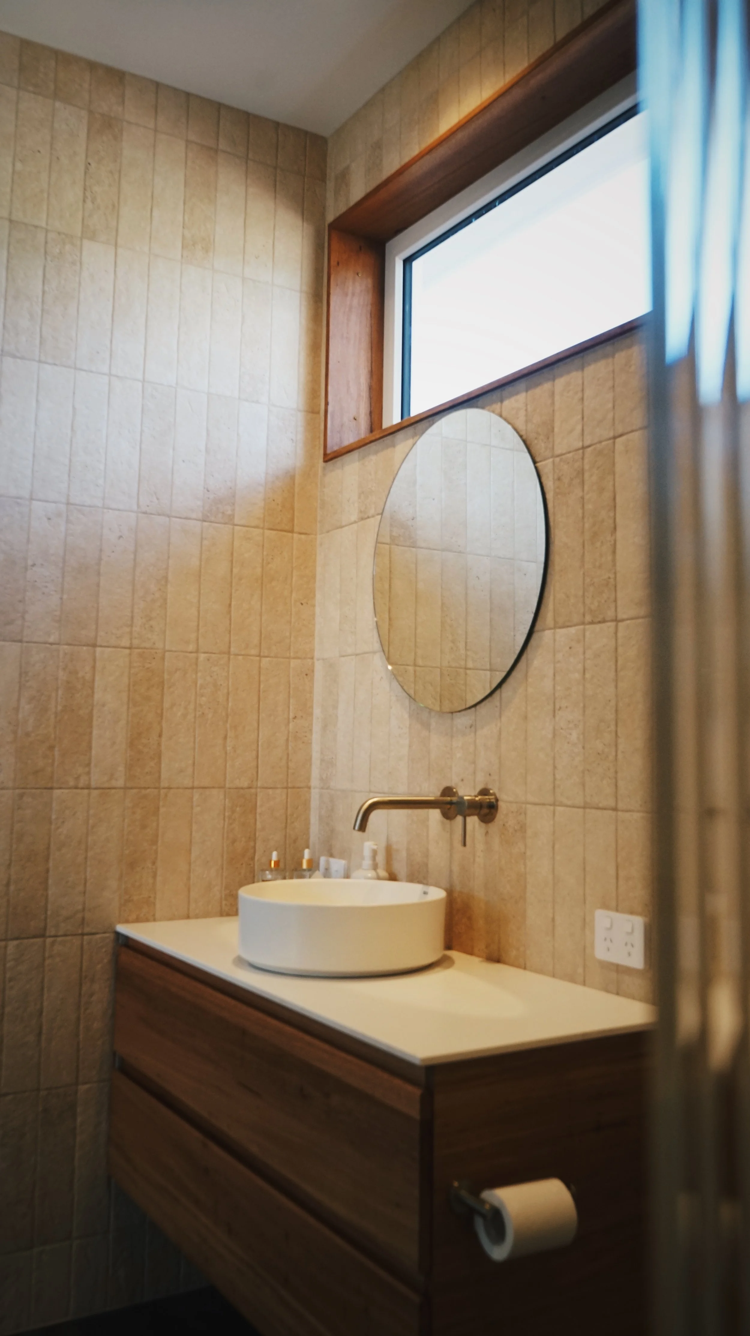 A bathroom with beige tiled walls, a round white vessel sink on a wooden vanity, a wall-mounted gold faucet, a round mirror above the sink, small bottles on the vanity, a window above the mirror with a wooden trim, and a white electrical outlet next 
