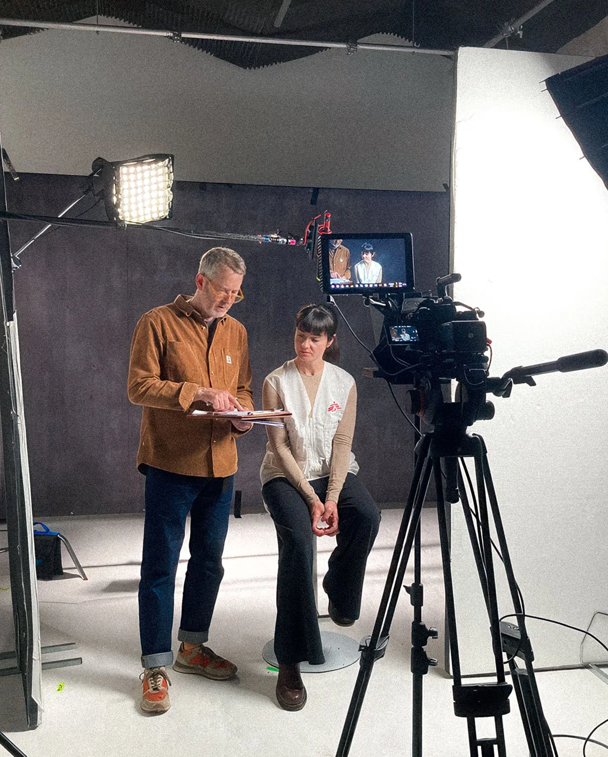 A man and a woman are in a studio with professional lighting and a black background. The man is standing and holding a notebook, looking down at it. The woman is sitting on a stool, looking at the man. A camera on a tripod is recording them, and a monitor displays their image, indicating they are filming a video or conducting an interview.