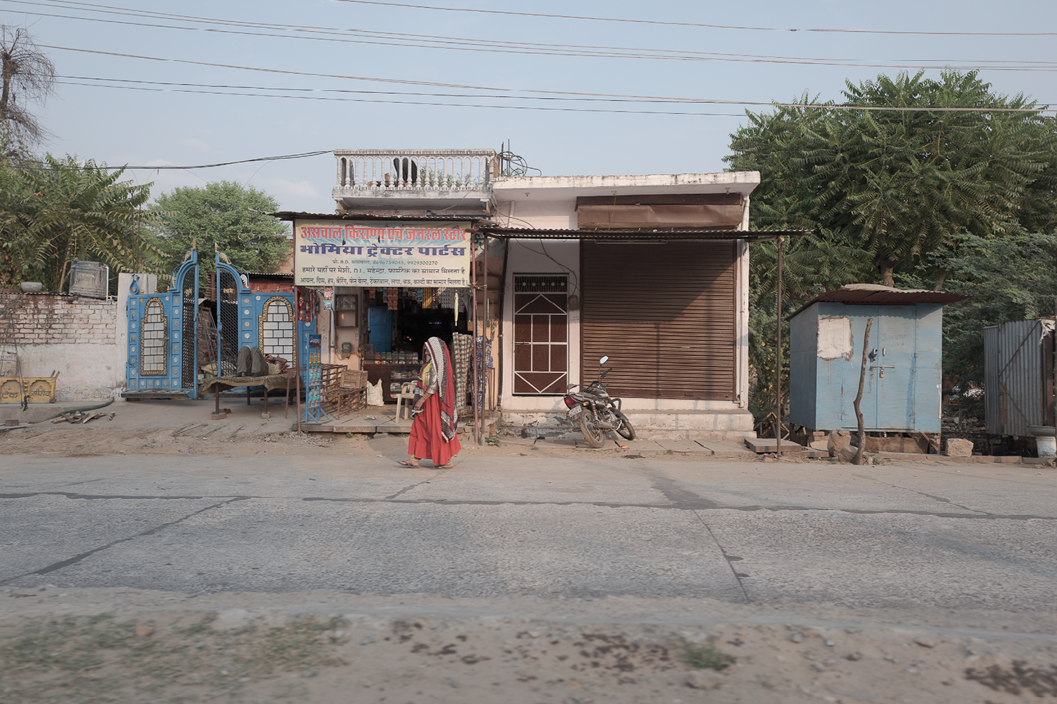 Rue avec une petite boutique et une femme en sari rouge marchant devant, moto garée devant la boutique, arbre vert en arrière-plan, ciel clair.