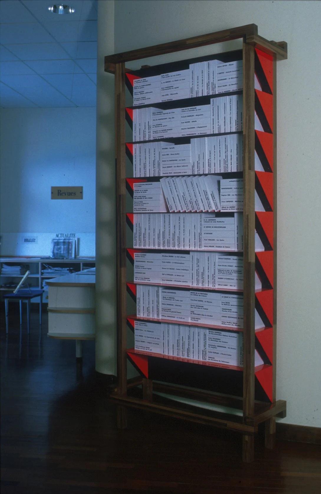 Un tableau d'affichage avec des billets ou des programmes, situé dans un bâtiment, probablement dans une bibliothèque ou un centre culturel, avec un fond montrant une salle avec des étagères et des affiches.