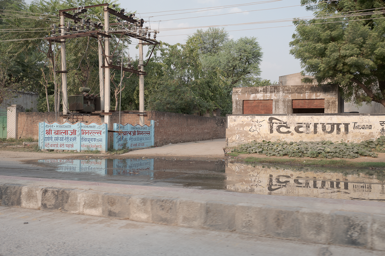 Une vieille structure électrique avec des poteaux et des fils électriques, un mur en briques et des panneaux publicitaires en langue hindi, avec des arbres en arrière-plan et une route en premier plan reflétée dans une flaque d'eau.