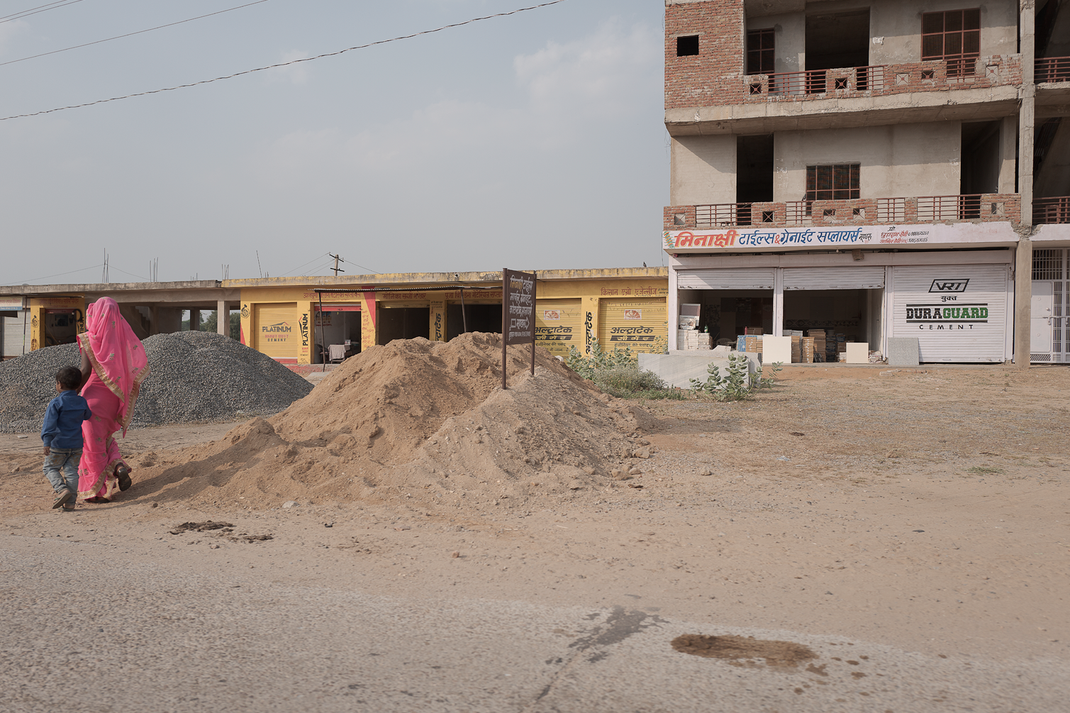 Deux enfants, une fille en sari rose et un garçon en chemise bleue, marchent à côté de tas de terre et de gravier sur un chantier, avec des bâtiments en construction et des boutiques fermées en arrière-plan.
