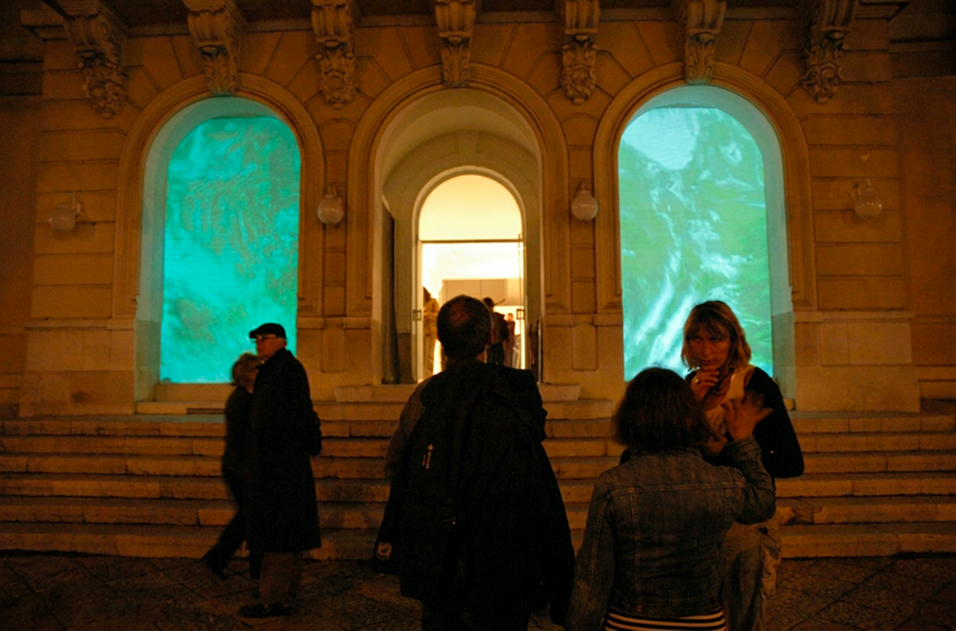 Groupes de personnes devant un bâtiment avec deux grandes fenêtres projetant des images bleues et vertes. L'atmosphère est nocturne.