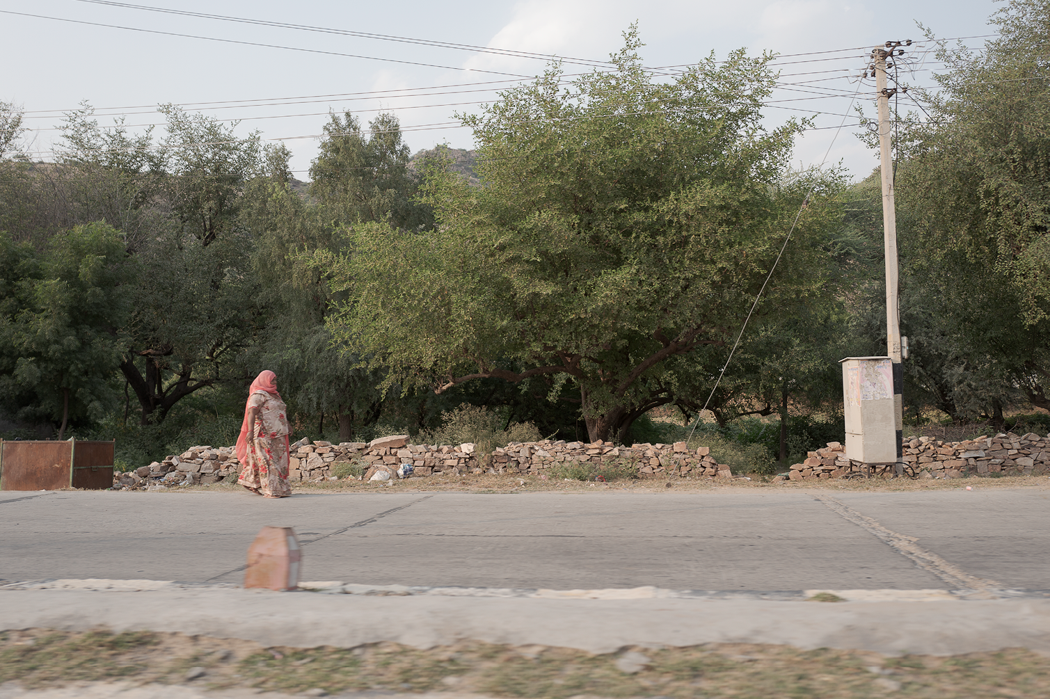 Une femme portant une robe longue à motifs et un voile rose marchant sur le trottoir, avec des arbres verts derrière un mur en pierres et une ligne électrique.