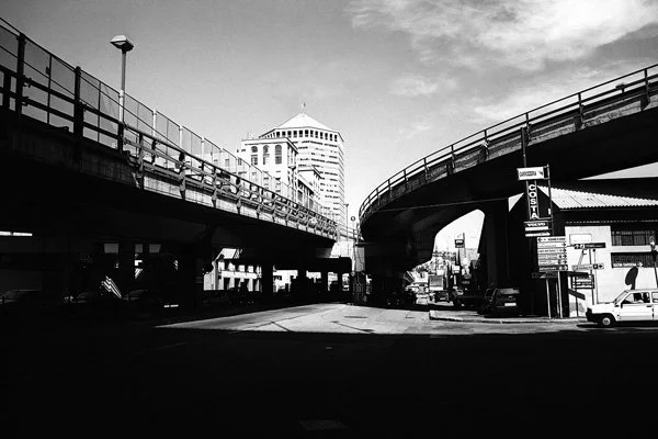 Vue en noir et blanc d'une intersection urbaine avec un pont élevé et un grand bâtiment en arrière-plan.