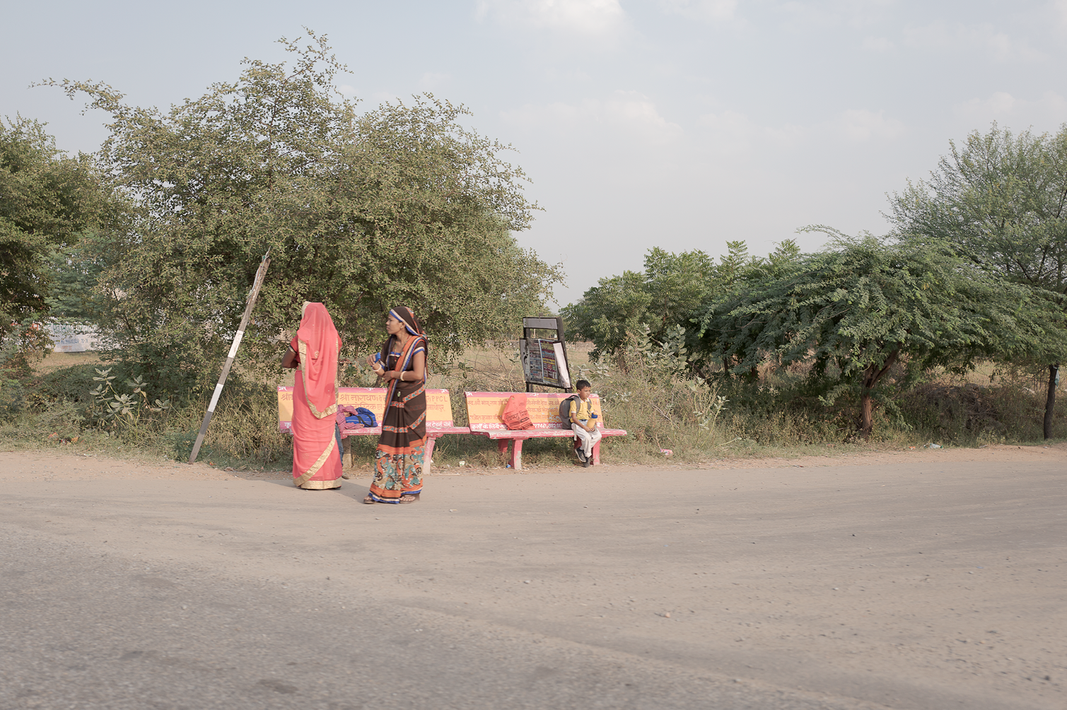 Deux femmes en sari, une assise sur un banc et une autre debout, près de plusieurs arbres et un homme avec un sac à dos qui s'assoit sur un banc peint en rose avec des arbres derrière, dans un paysage rural.