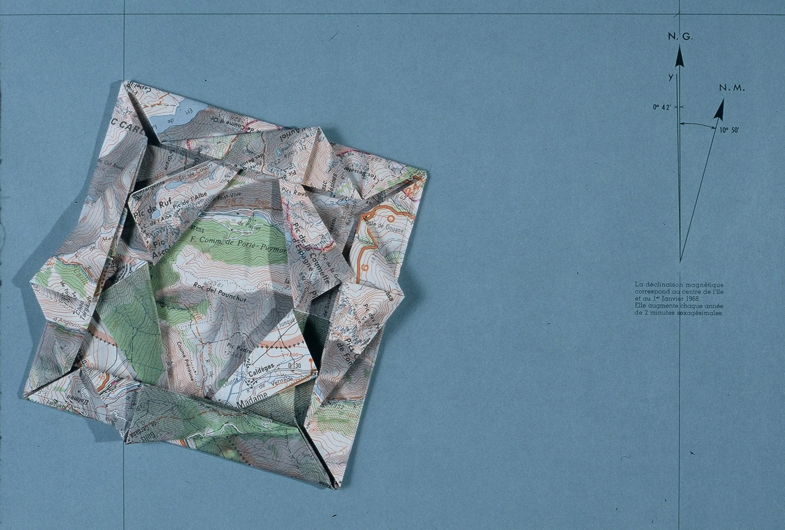 Une carte topographique pliée ressemblant à un papillon, posée sur une surface bleue. La carte montre des détails géographiques et une déclaration en français en bas à droite.