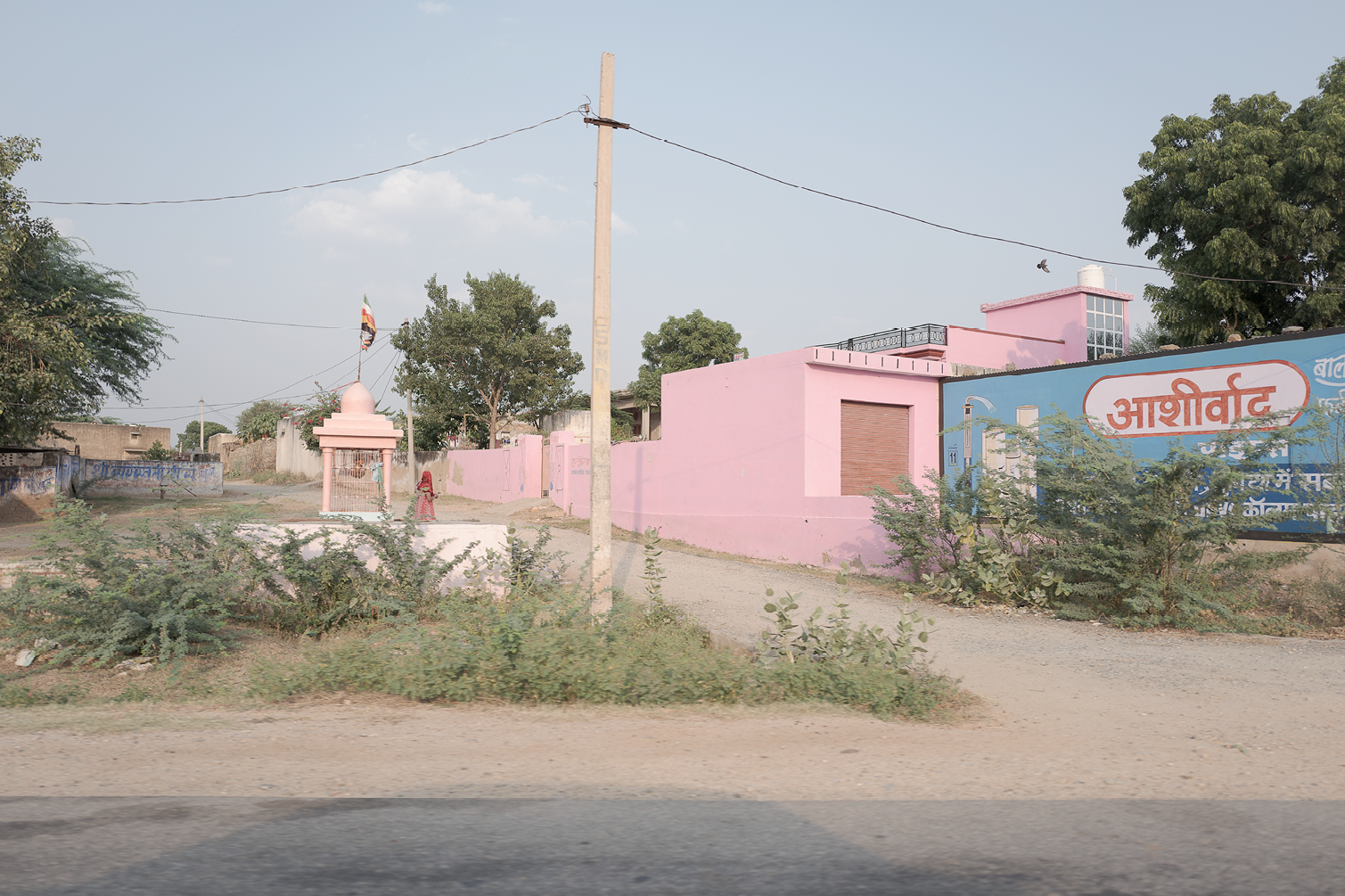 Vue d'une rue rurale avec un mur rose, un petit temple, quelques arbres et une femme portant un sari rouge. Signage publicitaire en hindi.