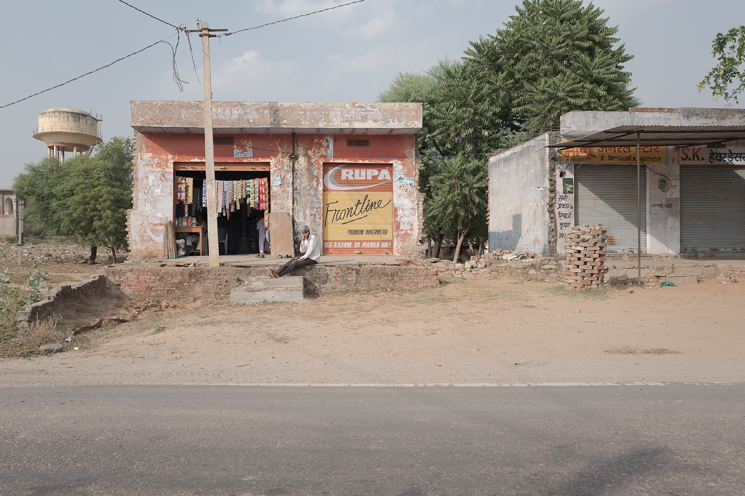 Une petite boutique en ruine avec un homme assis devant, une chaîne de vêtements suspendue à l'entrée, et une affiche peinte sur la porte avec le nom "Rupa". Le bâtiment est entouré d'arbres et d'un ciel nuageux, avec une route asphaltée au premier p