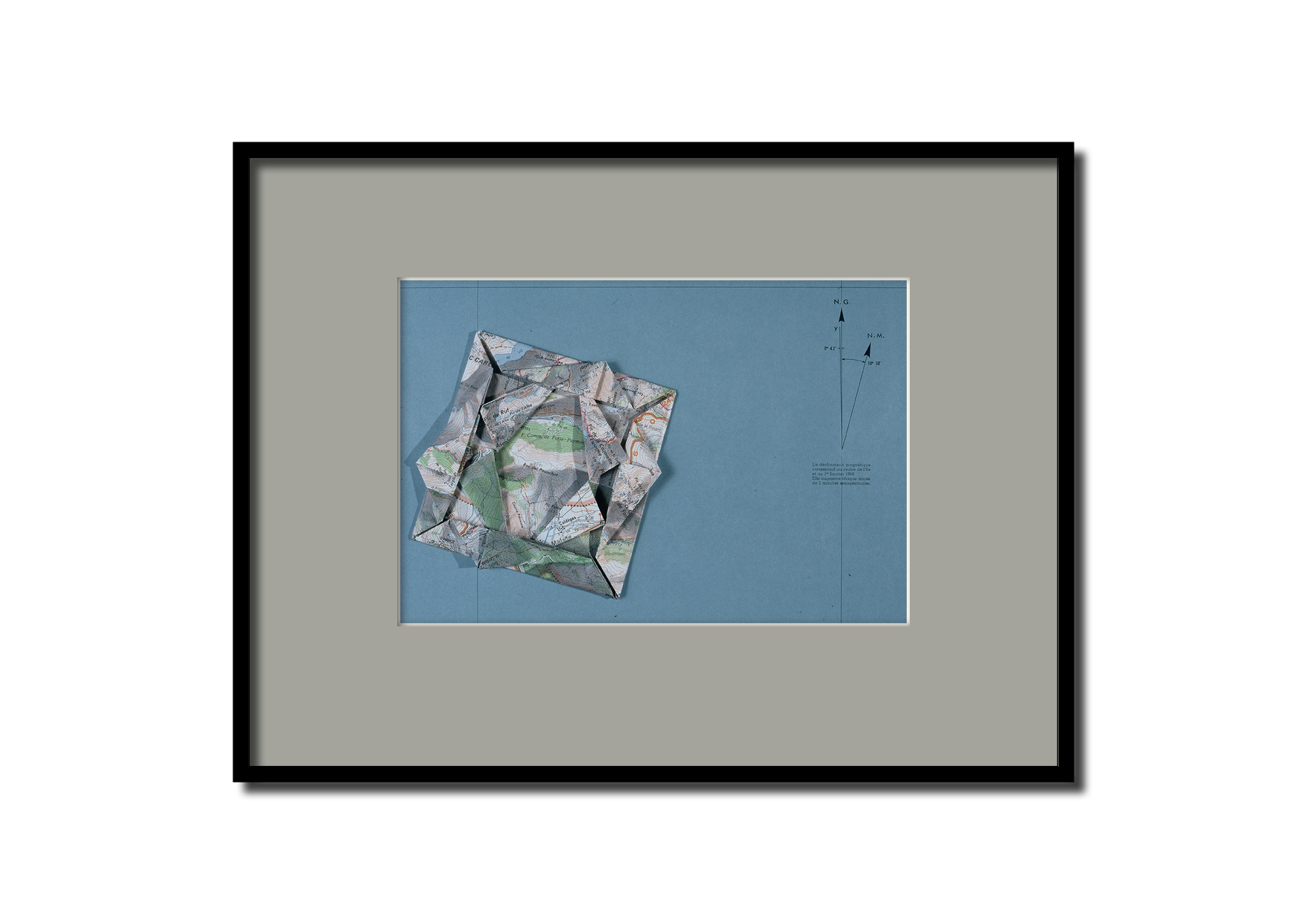 Une carte pliée en trois dimensions posée sur une surface bleue, avec un compas et des annotations de direction, montrant des détails topographiques et géographiques.