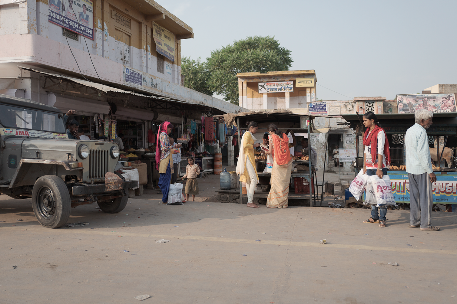 Une scène de marché en Inde avec des vendeurs, des acheteurs et une Jeep ancienne sur une route poussiéreuse.