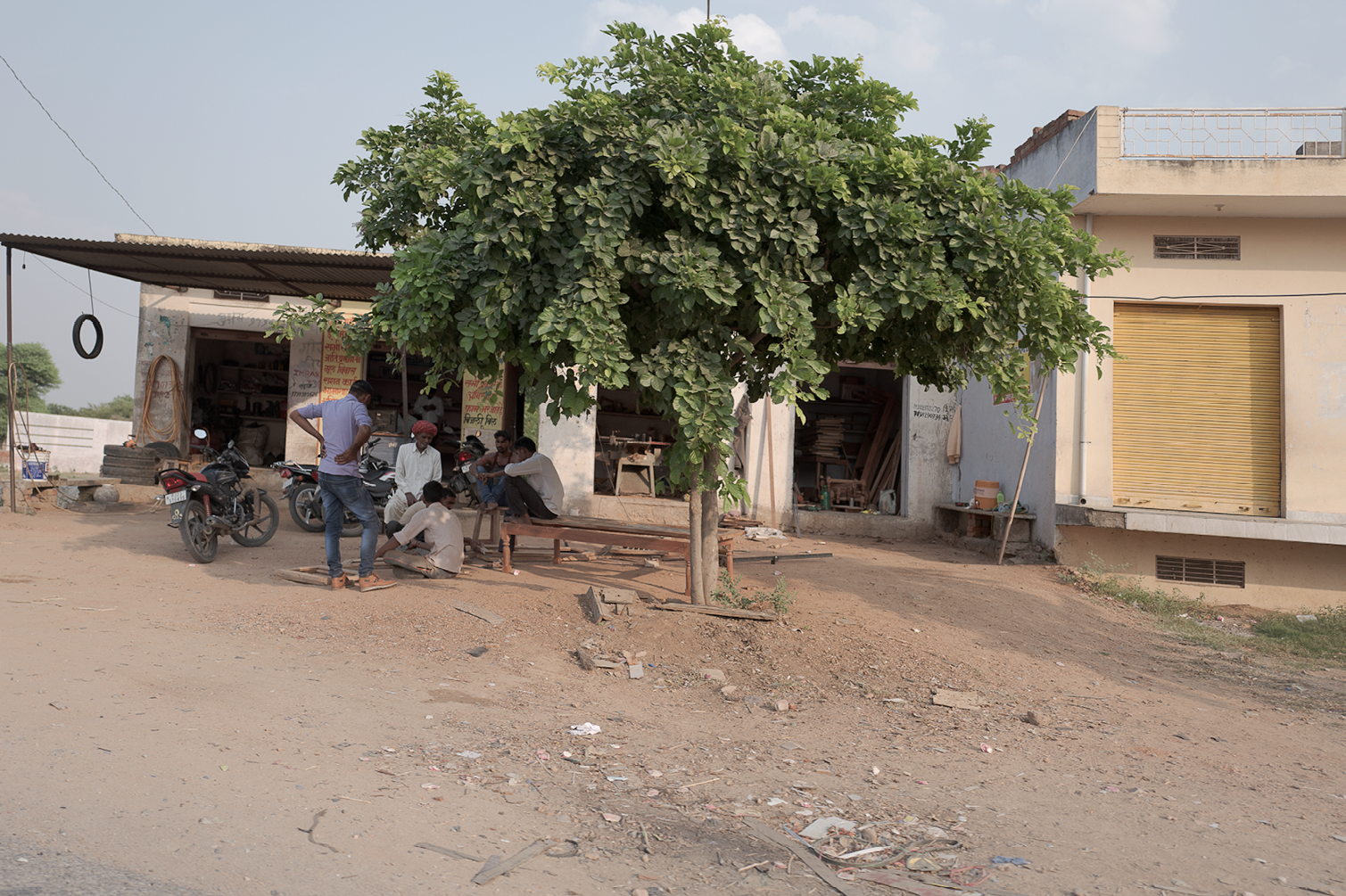 Groupe de personnes assises et discutant sous un arbre au bord d'une route poussiéreuse, avec des motos garées devant un atelier ou magasin.