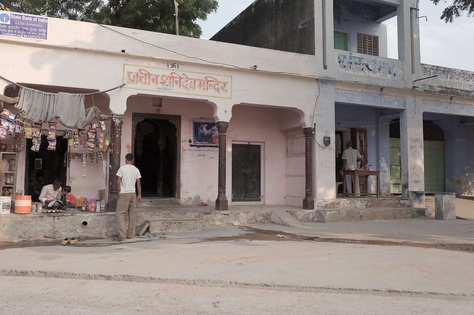 Une façade de bâtiment en Inde avec des boutiques et des personnes assises ou en train de faire des activités à l'extérieur. Il y a un panneau indiquant une banque et une écriture en hindi.