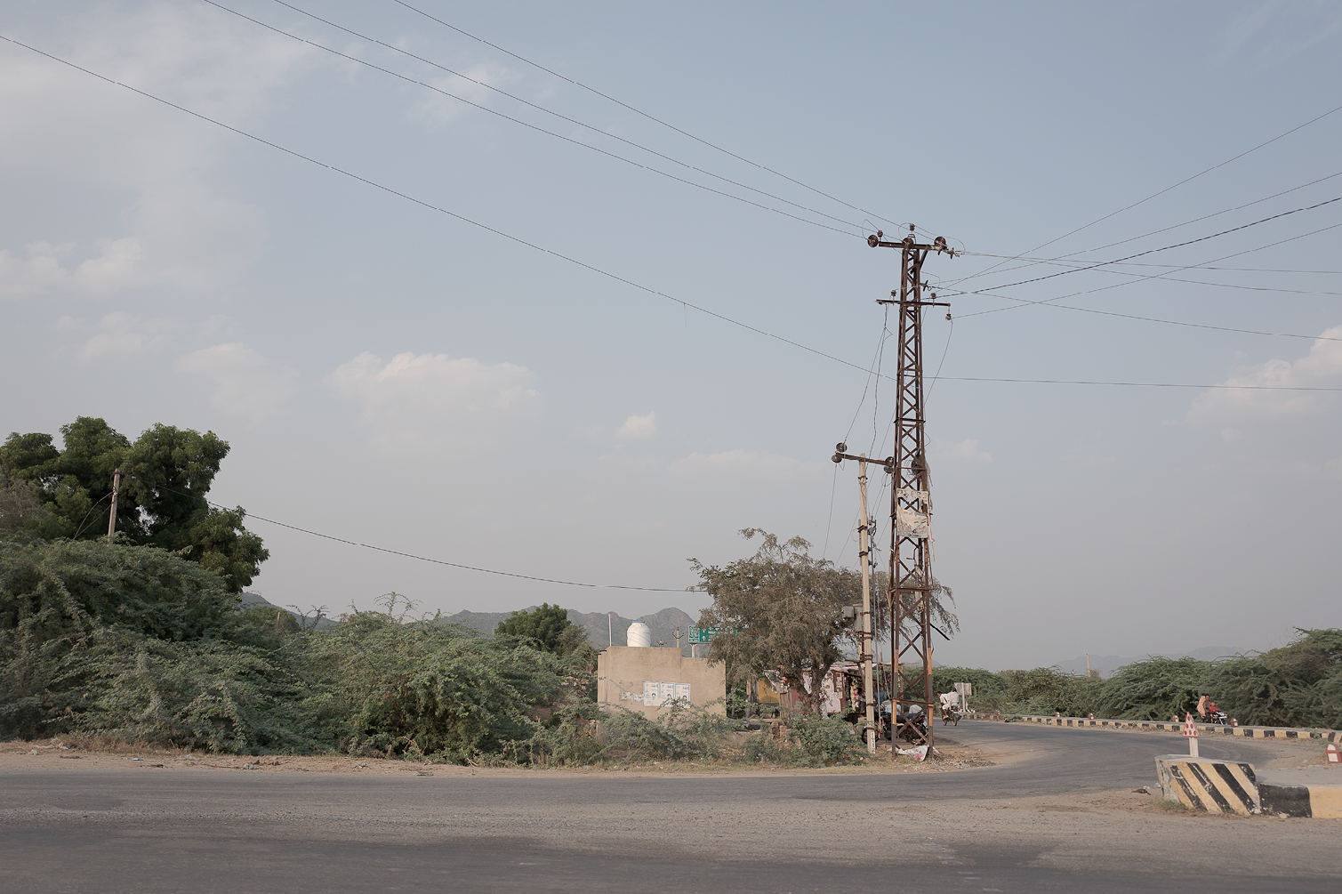 Un paysage rural avec une route, des arbres, un poteau électrique en métal rouillé, quelques bâtiments en arrière-plan et des cyclistes sur la route, ciel partiellement nuageux.