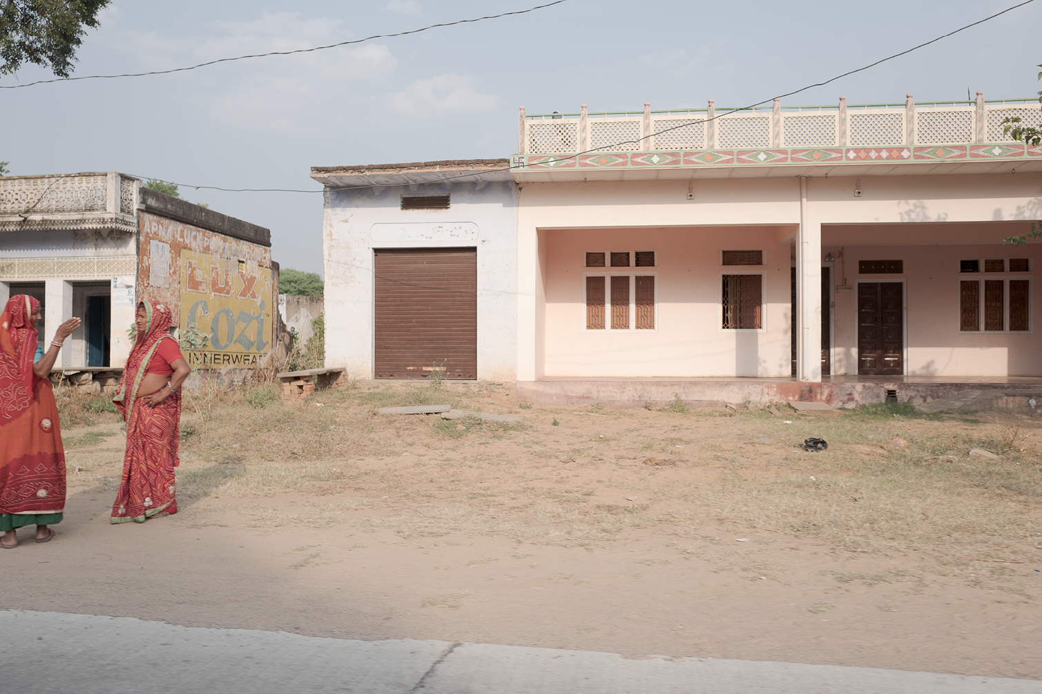 Deux femmes vêtues de saris rouges discutent devant des bâtiments simples, l'un avec un mur peint et un autre en construction, dans un environnement urbain en Inde.