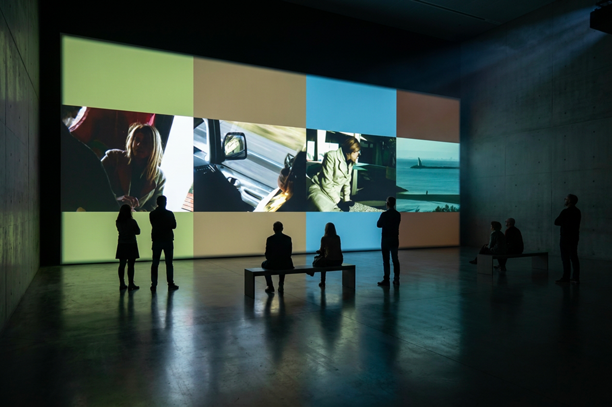 Une salle d'exposition sombre avec un grand mur de projection affichant plusieurs images de personnes et un avion en mer, avec quelques personnes debout ou assises regardant l'écran.