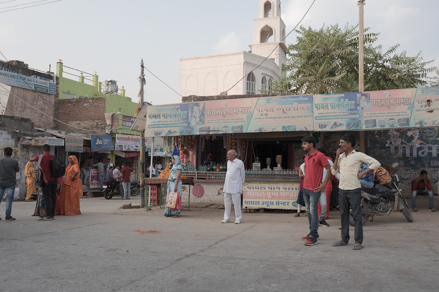 Une scène de rue avec plusieurs personnes devant un magasin de produits divers, dont des bouteilles en plastique. Certains hommes discutent, une femme porte un sac, d'autres regardent autour. En arrière-plan, un bâtiment avec une église ou une mosqué