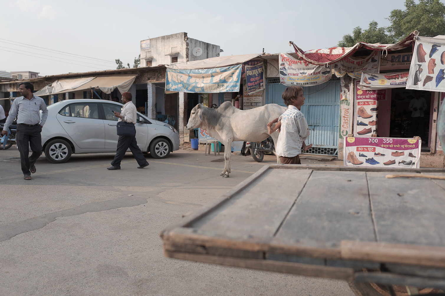 Une rue animée avec des personnes marchant, une vache, une voiture, et des boutiques vendant des chaussures.