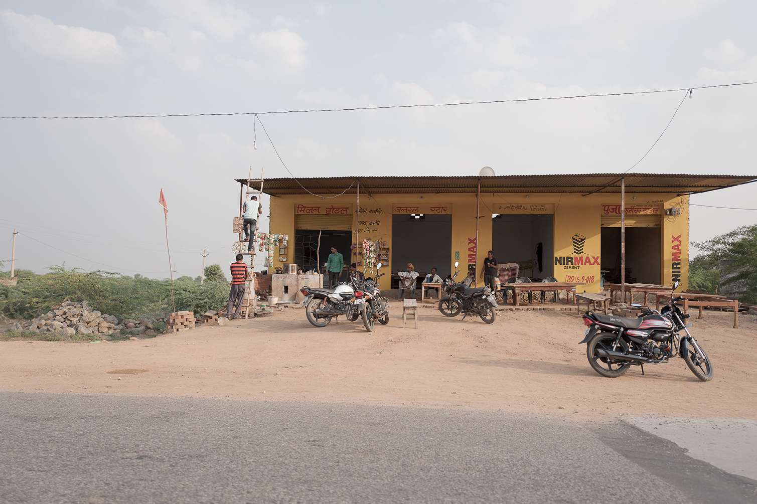 Une petite boutique au bord de la route avec des motos garées devant, quelques personnes assises ou debout, un homme grimpe un échafaudage, et le décor est rural avec des arbres et un ciel partiellement nuageux.