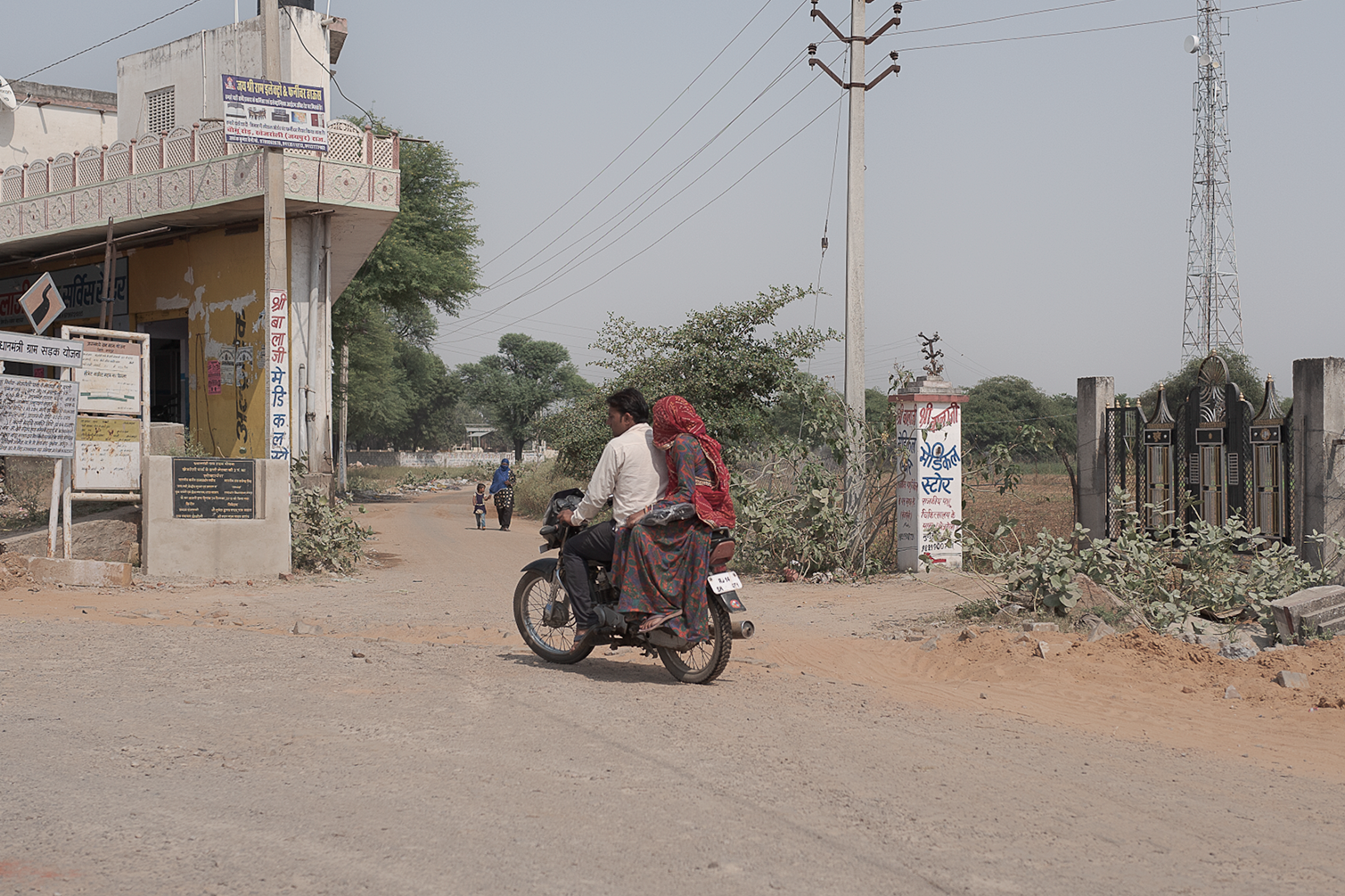 Deux personnes, un homme et une femme à l'arrière, circulent en scooter dans une rue de village ou de petite ville, avec un bâtiment, des arbres, des poteaux électriques et un panneau publicitaire en hindi en arrière-plan.
