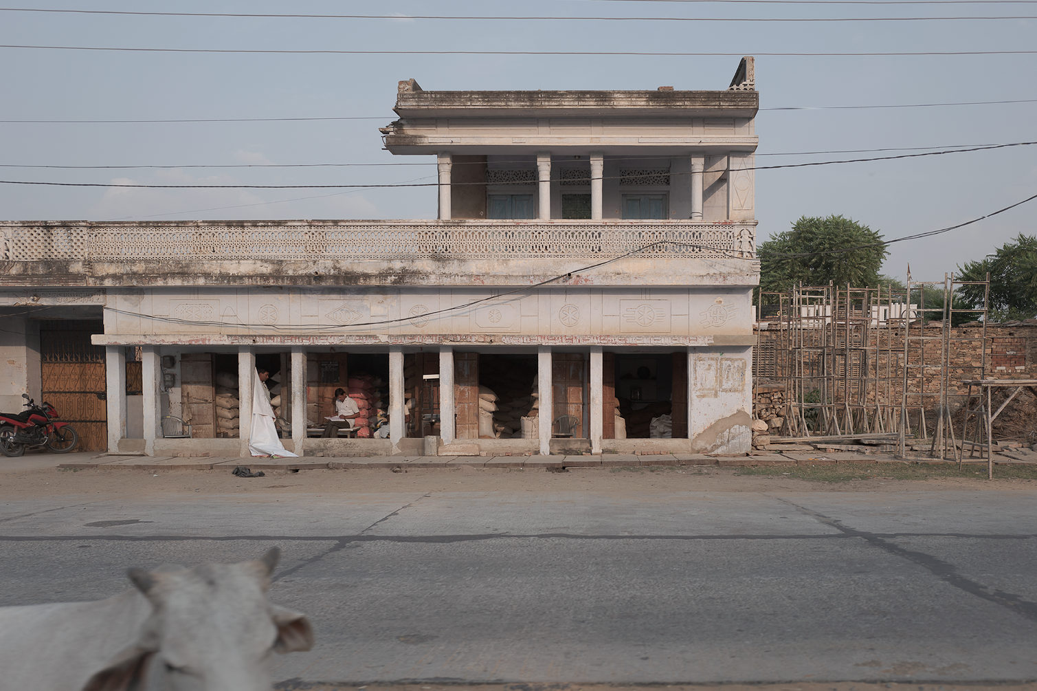 Une façade vieillie d'un bâtiment avec des colonnes, des sacs de marchandises à l'intérieur, un chien en premier plan et un mur en briques à droite, dans un cadre rural ou ouvrier.