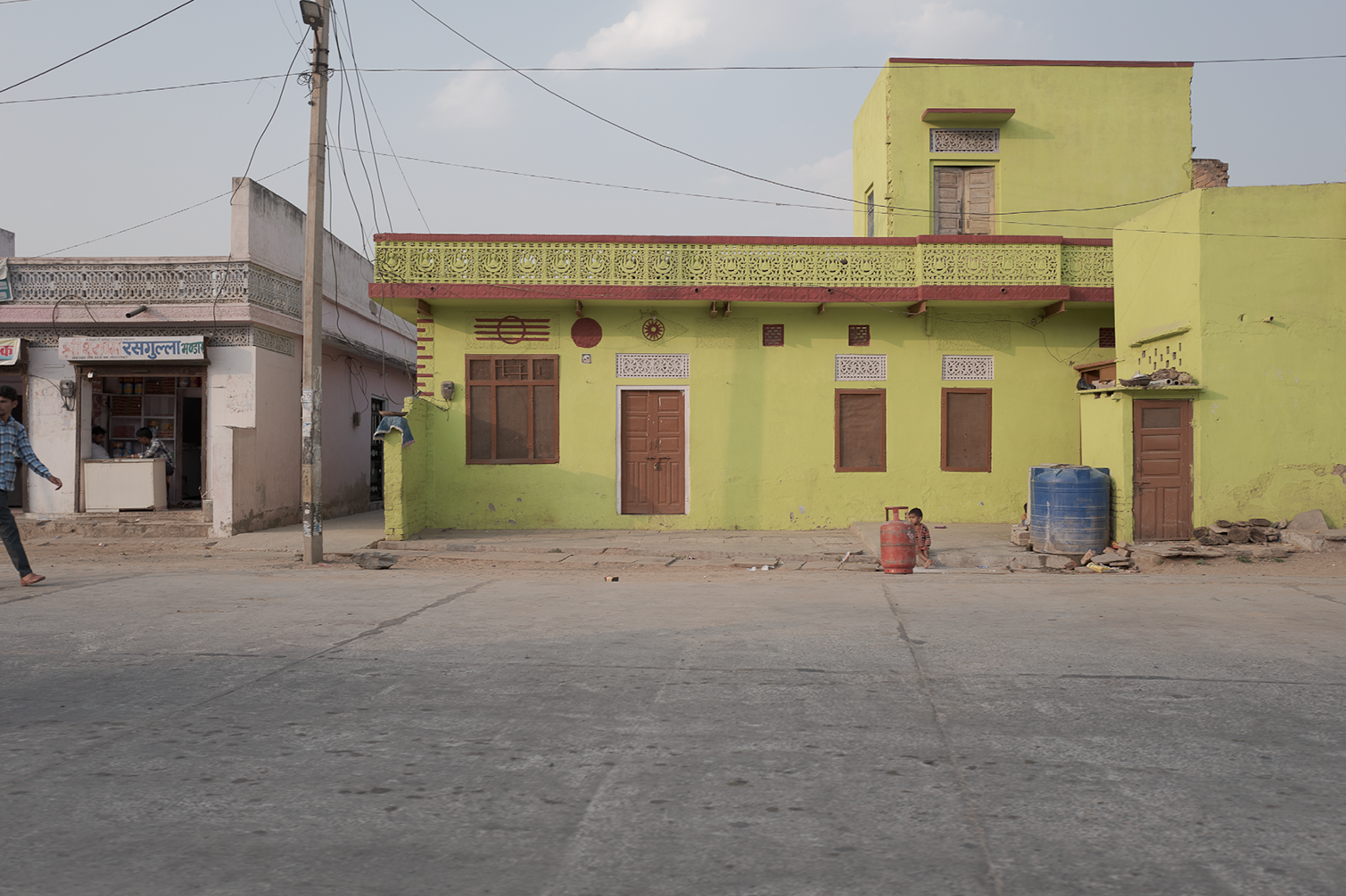 Une maison verte avec un balcon ornemental en haut, située sur une rue pavée, avec deux enfants, un d'entre eux assis et l'autre marchant, et des bidons en plastique à l’extérieur.