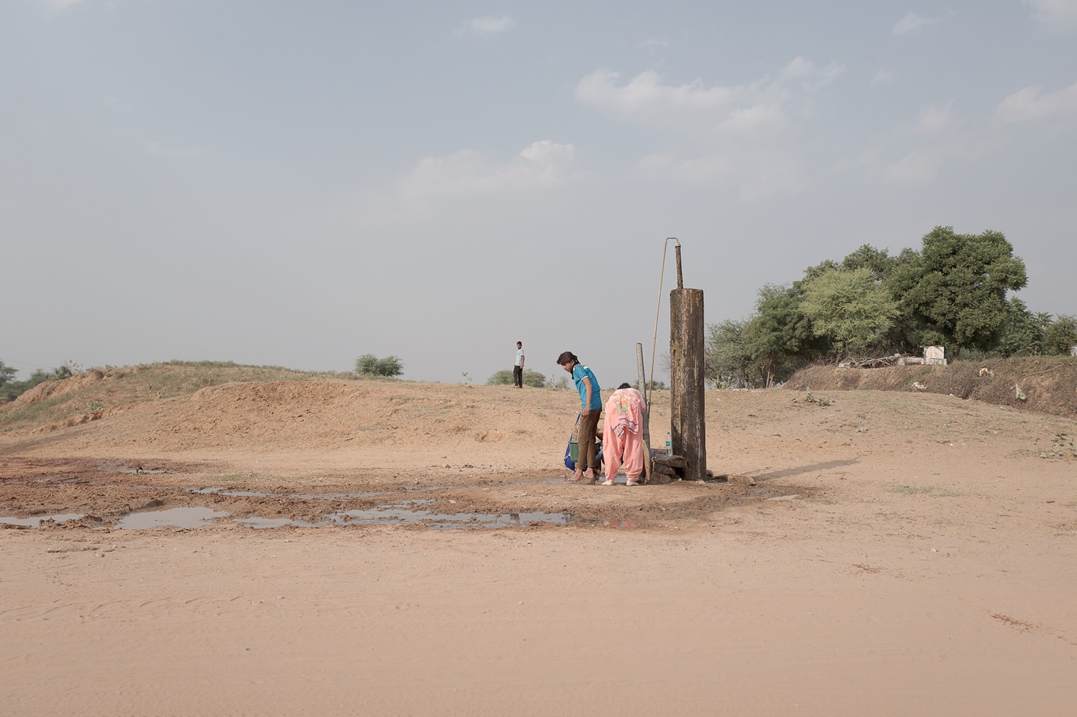 Groupe de personnes près d'une pompe à eau en plein air, dans un paysage désertique avec quelques arbres en arrière-plan.