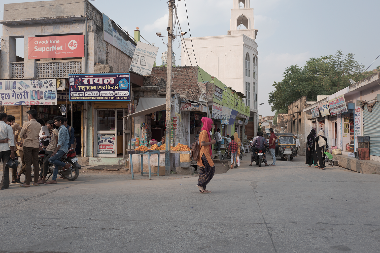 Vue d'une rue animée en Inde avec des marchands, des passants et des véhicules, incluant une mosquée blanche en arrière-plan.