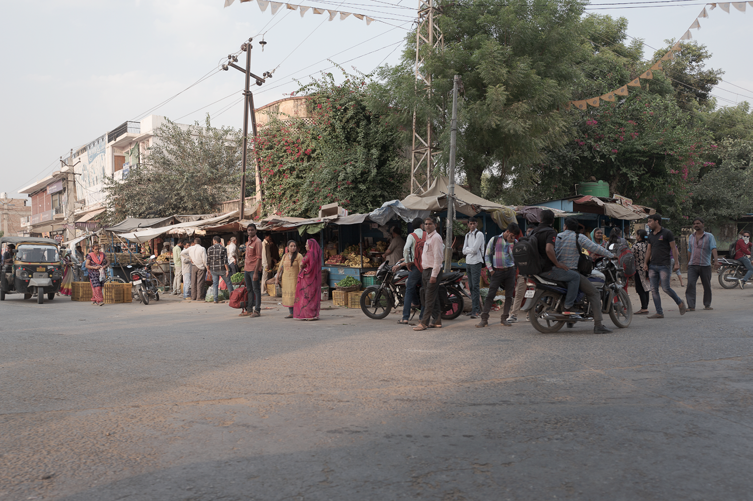 Un marché en plein air avec des vendeurs et des acheteurs, y compris des personnes sur des motos, dans une ville en Inde, avec des arbres, des échoppes, et des boules de papier suspendues en décoration.