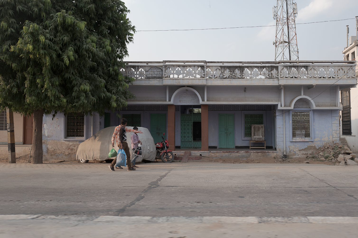 Vue de la façade d'une maison ancienne avec deux portes vertes et fenêtres grillagées, un arbre sur la gauche, deux personnes marchant sur la route devant la maison, une moto garée et un air conditionné devant la maison.