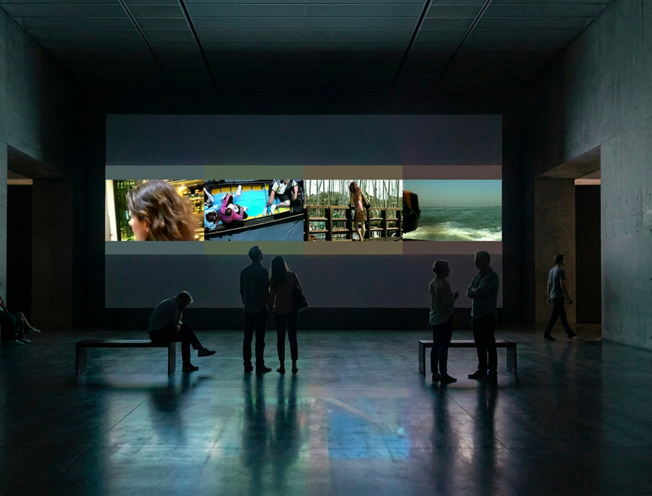 Salle d'exposition sombre avec un groupe de personnes regardant une grande œuvre d'art ou projection video, plusieurs silhouettes sont visibles silhouettées contre l'écran.