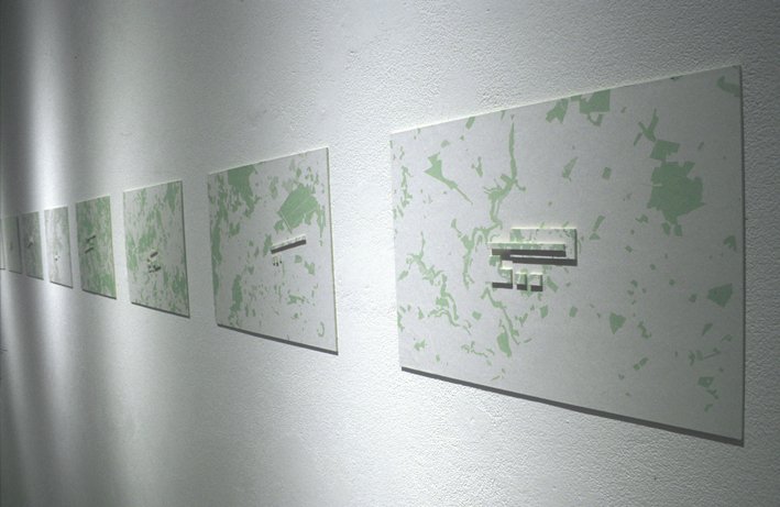 Une série de tableaux abstraits avec un fond blanc et des motifs verts, accrochés en ligne sur un mur blanc.
