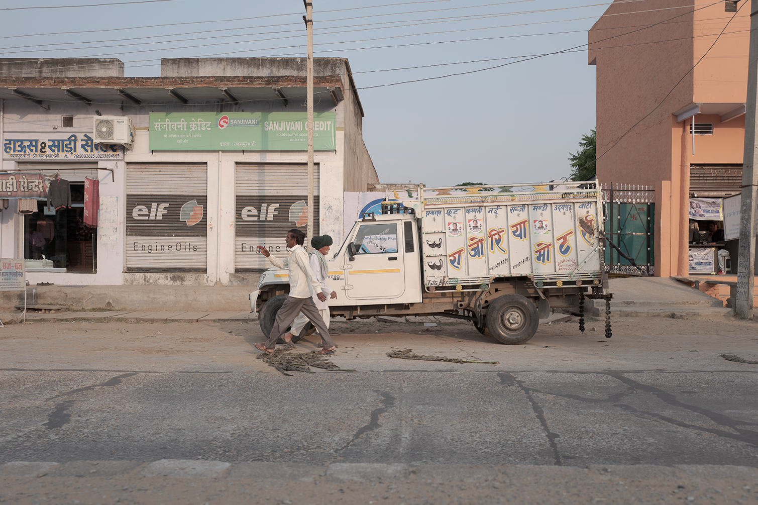 Deux personnes marchent côte à côte devant un camion blanc garé sur la route d'une ville. En arrière-plan, il y a des magasins avec des enseignes, dont une pour les huiles moteur Elf, et un bâtiment en pierre de couleur beige. La route est en mauvais