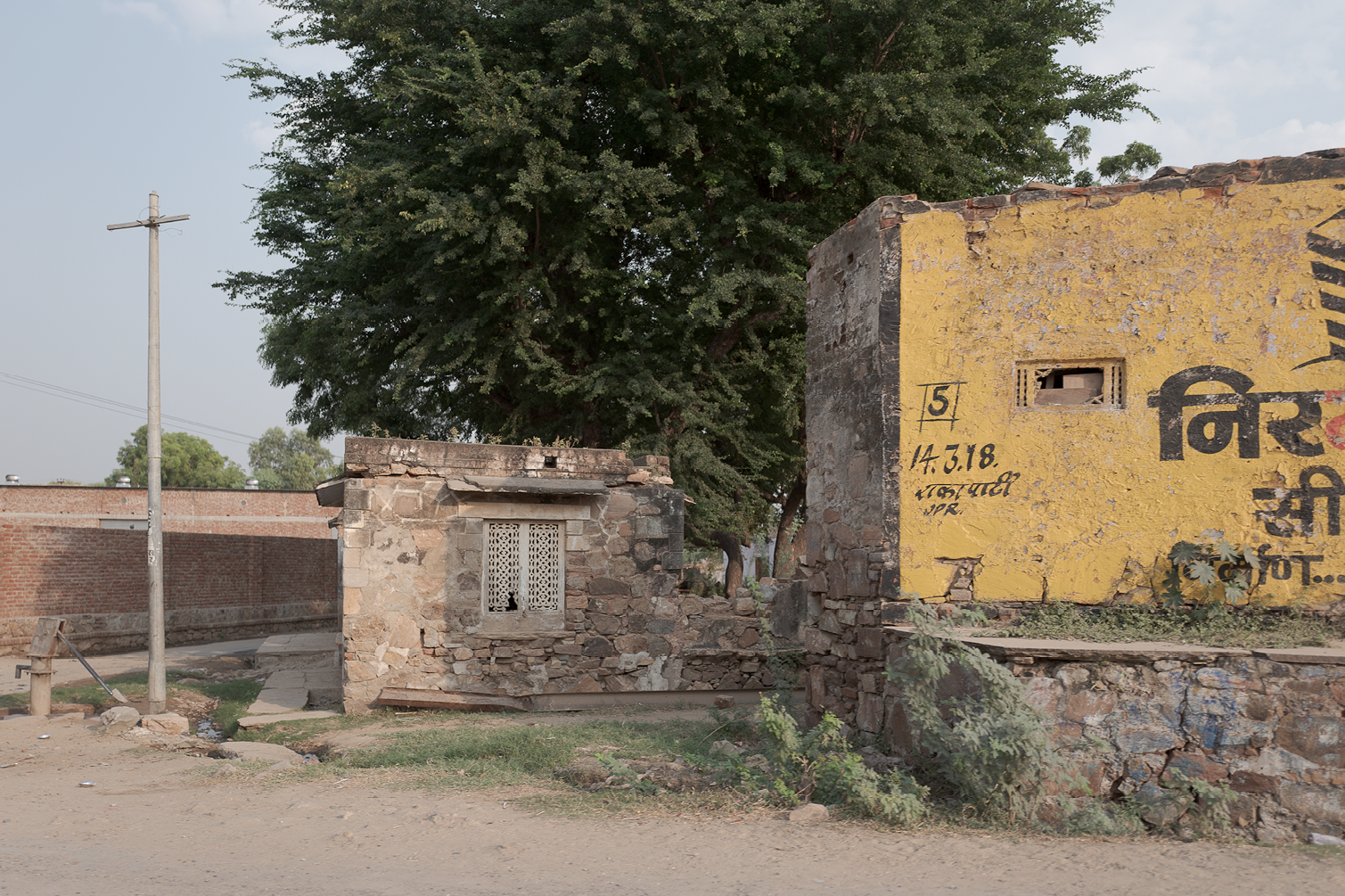 Ancienne maison en pierre avec fenêtre en métal, mur en pierre et mur en pierre peint en jaune avec inscriptions en hindi, grand arbre en arrière-plan, poteau électrique à gauche, sol en terre battue avec quelques plantes