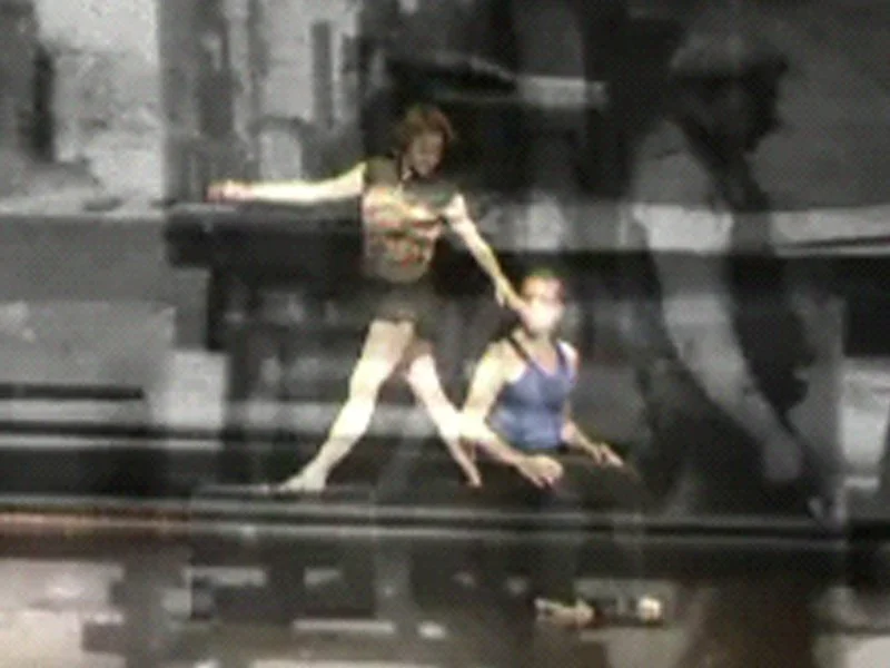 Une scène dans un auditorium ou un studio, avec deux danseuses, une assise et une debout, faisant une performance de danse. La scène est sombre avec un fond industriel.