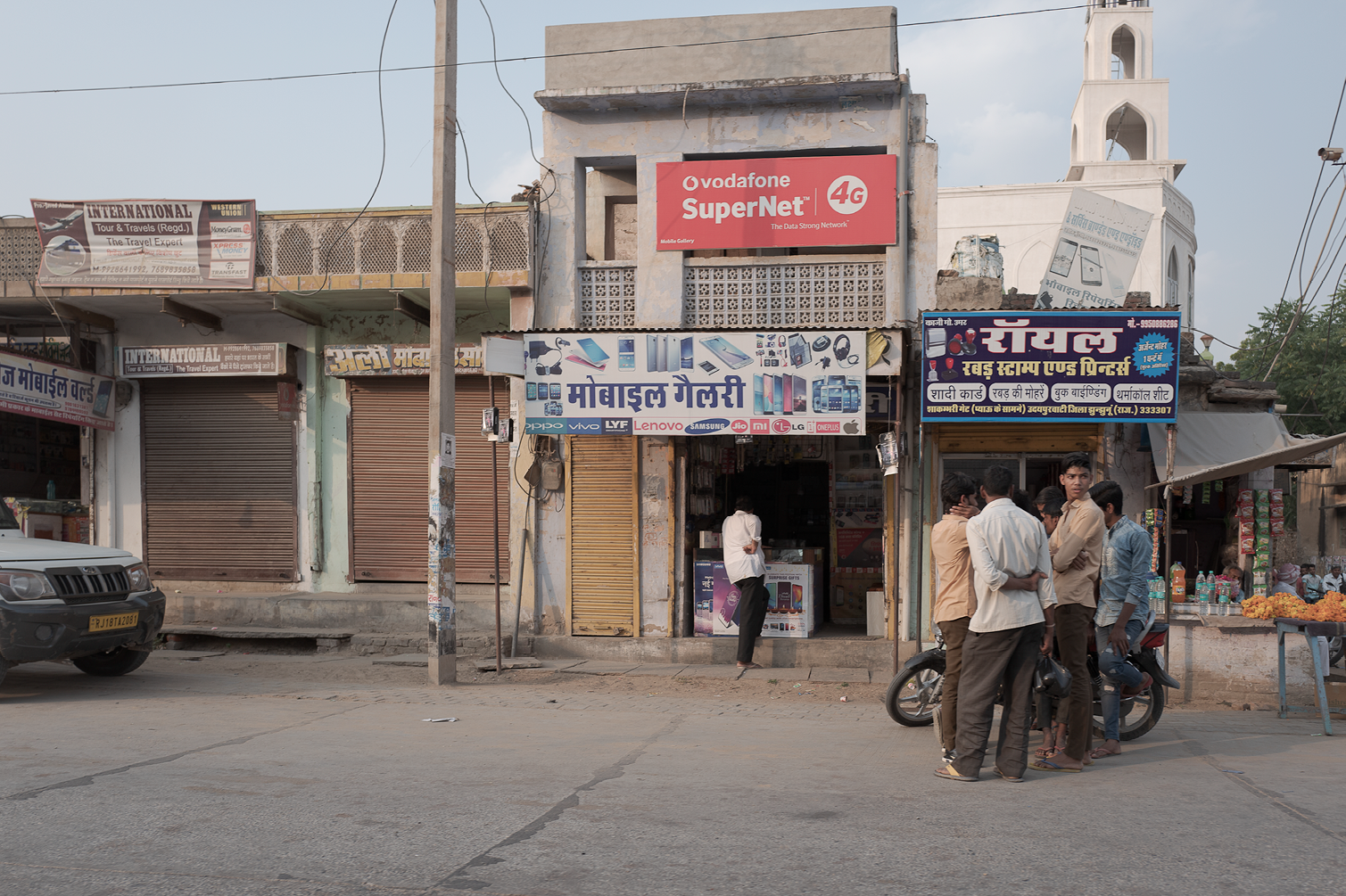 Une rue indienne avec des boutiques fermées et un groupe de personnes discutant devant un stand. Il y a aussi une boutique de téléphones mobiles ouverte au centre.