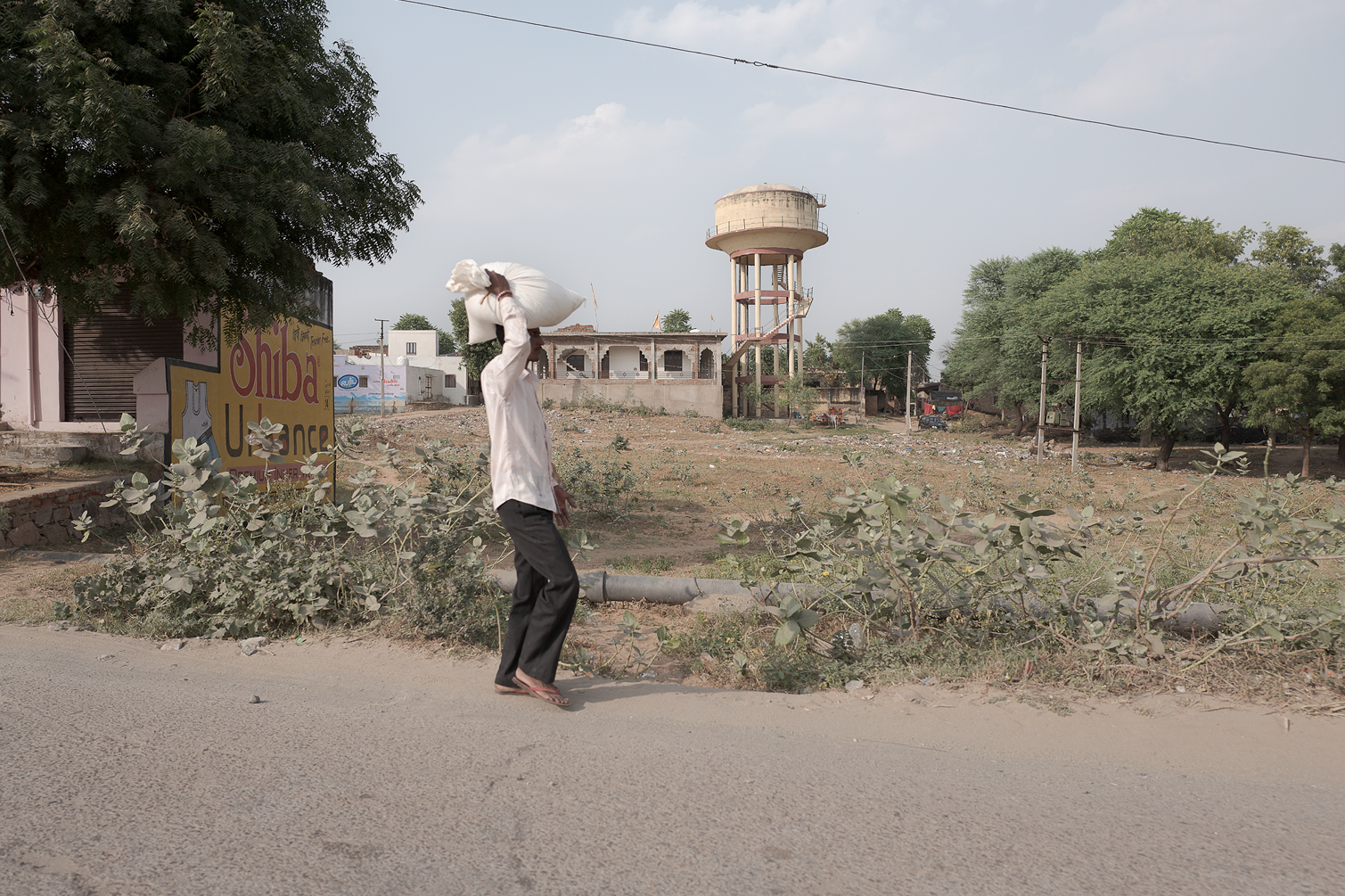 Un homme portant un sac blanc sur l'épaule marche le long d'une route poussiéreuse, avec des arbres et des bâtiments en arrière-plan, dans un paysage semi-urbain.