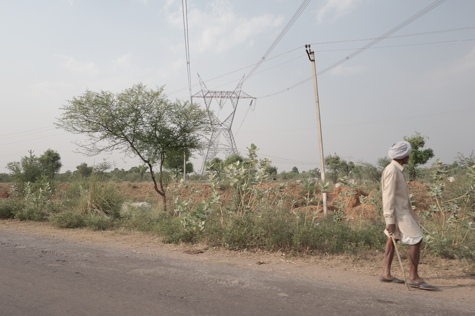Un homme en vêtements traditionnels marche le long d'une route en terre dans une zone aride avec quelques arbres et végétation, à côté de lignes électriques et un pylône dans le paysage