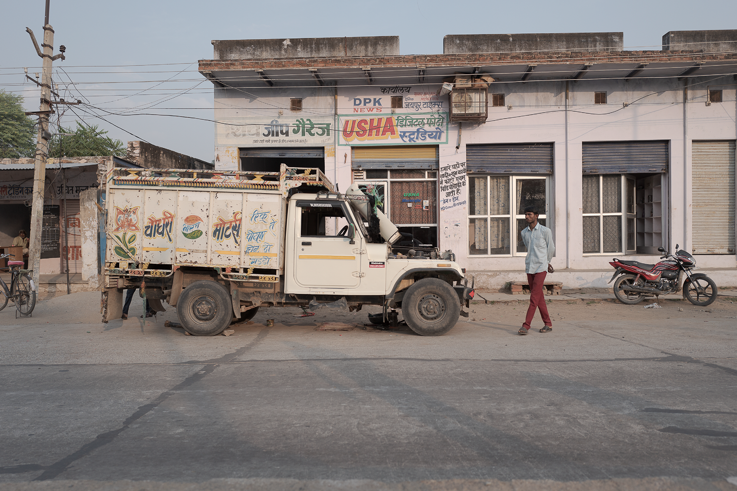 Une rue en Inde avec une camionnette blanche décorée, un homme marche à côté, et un bâtiment avec des panneaux publicitaires et des fenêtres. Une moto est stationnée près du bâtiment.