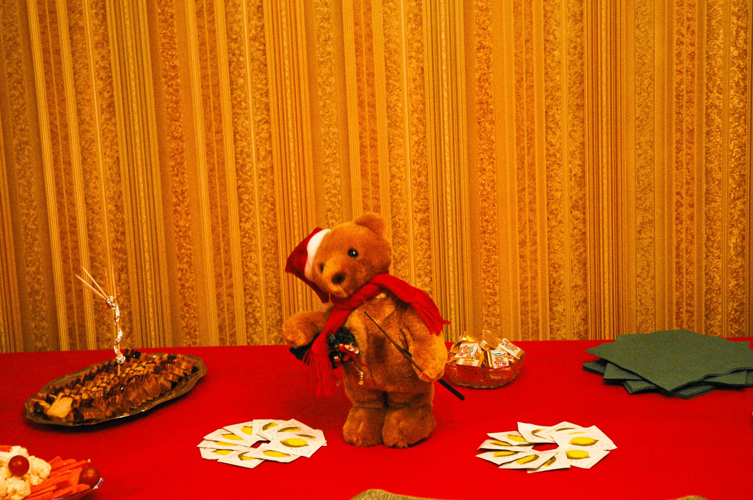 Un ourson en peluche portant un chapeau rouge et une écharpe rouge est placé sur une table rouge, entouré de cartes à jouer avec un motif de citron. À côté, il y a un plat de gâteau, quelques papiers verts pliés, et un panier de bonbons. En arrière-plan, un mur avec un motif de rayures verticales beiges et marron.