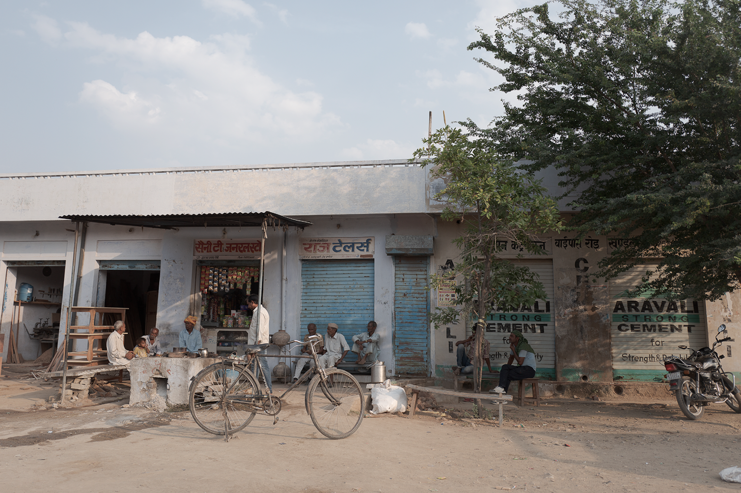 Une rue calme avec des bâtiments simples, des personnes assises, un vélo, et une moto, dans un environnement rural ou semi-urbain en Inde.