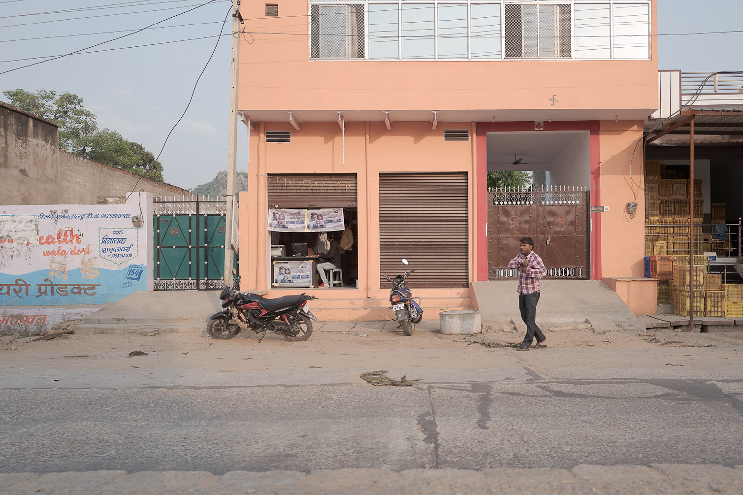 Une rue simple avec une bâtiment à façade rose, une moto et un scooter garés devant, une personne marchant en portant une bouteille, et un petit espace avec un ordinateur où une autre personne travaille, avec des fenêtres et un portail métallique.