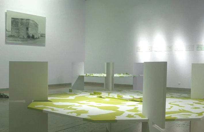 Une pièce d'exposition avec des structures en forme de tables et des panneaux, une peinture murale en noir et blanc, murs blancs et lumière naturelle